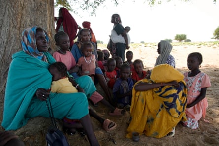 A group of scared women and children sit around the base of a tree; one woman prays using rosary beads, while holding a child close to her.